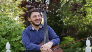 Harry Grossman, wearing a blue sweater with a button-down underneath, poses smiling with his bass in front of trees.