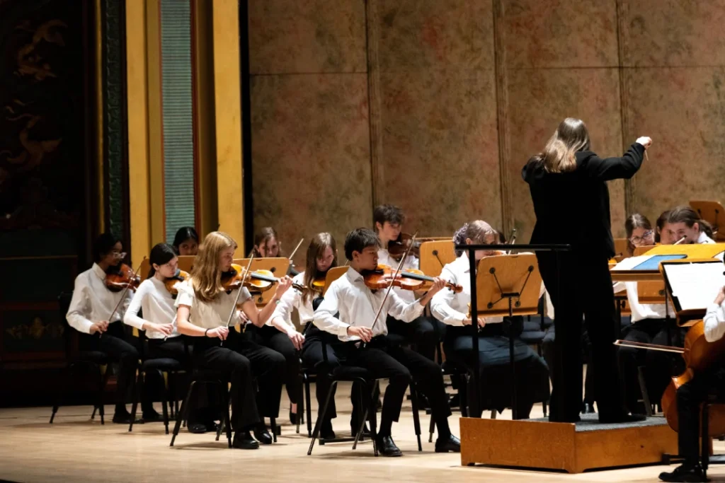 A conductor, dressed in all black, stands on the podium while conducting the students of Camerata Strings. Several rows of violinists, dressed in concert black and white, are visible.